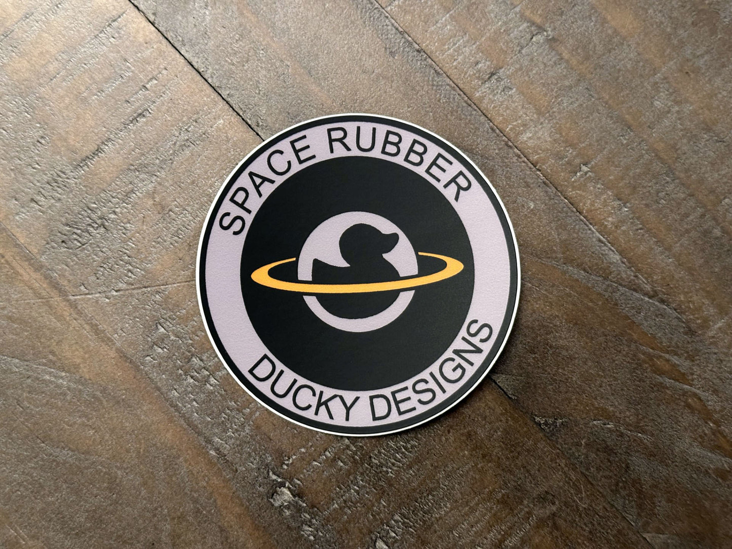 Space Rubber Ducky Designs decal sticker. 3" circle die cut vinyl decal. Matte finish and weather resistant. Features the company logo. 