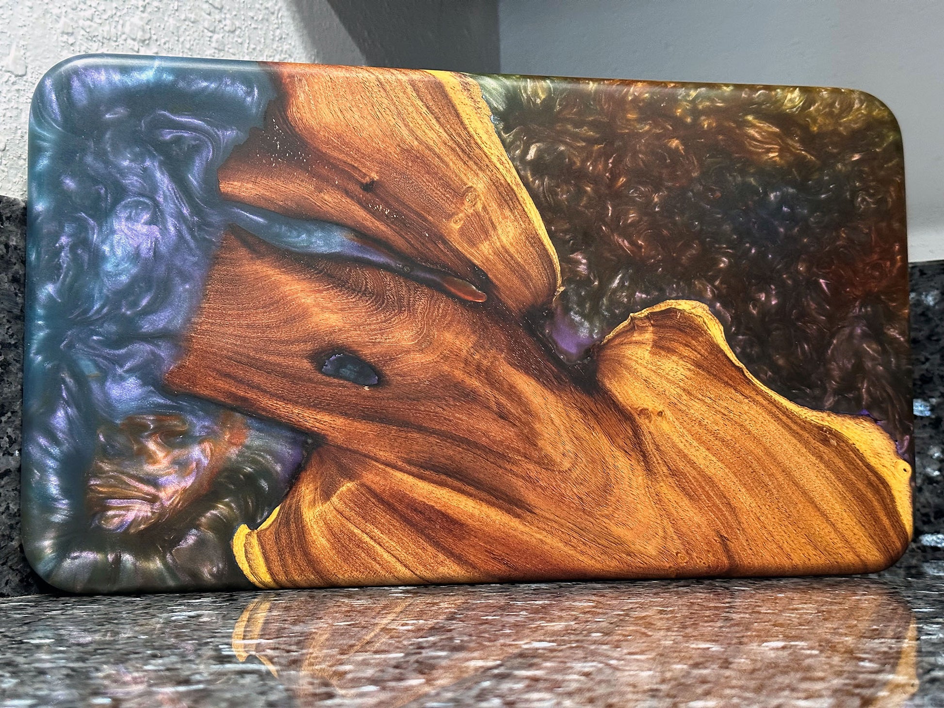The natural beauty of this live edge mesquite serving board is accented with a blast of color.  Dark blues and purples swirl together like a galactic nebula while a bubbling sea of color hypnotizes.   Food safe epoxy resin was used to create this design. Using knives or other sharp utensils on the epoxy resin surface may damage it and is not recommended.  Board dimensions:  17"L x 9.75"W x 0.75"H.  As a handcrafted product, please allow for approximately 1/4" variation in dimensions.