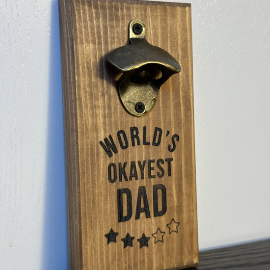 Custom magnetic bottle opener featuring a "World's Okayest Dad" theme. Each bottle opener is handmade and has hidden rare earth magnets to catch metal bottle caps. Wall-mounted or magnetic backing options. Made by Space Rubber Ducky Designs in Southern California, USA.